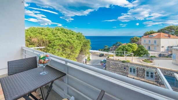 A balcony with a table and a glass of drink, overlooking a coastal town with buildings and a clear blue sky. - Apartment Luce Royal (Dubrovnik)