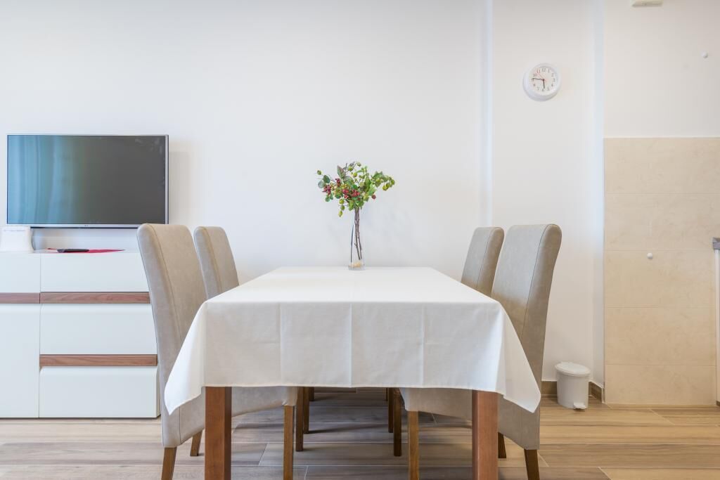 Ein Essbereich mit Tisch für zwei Personen, einem Blumenstrauß in einer Vase, einem Fernseher an der Wand und einer Uhr.
