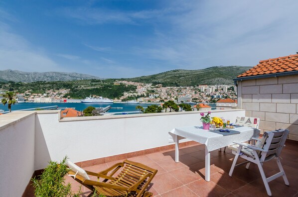 Une terrasse avec une table et des chaises, d’où l’on a vue sur une ville côtière et la mer.