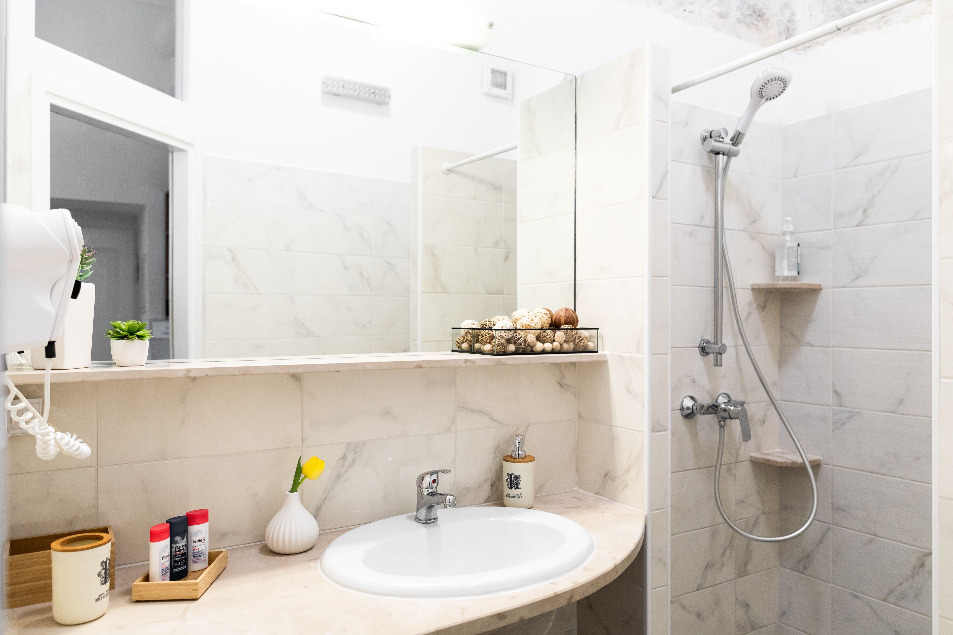 Un baño moderno con lavamanos blanco, espejo, ducha y estante con objetos decorativos.
