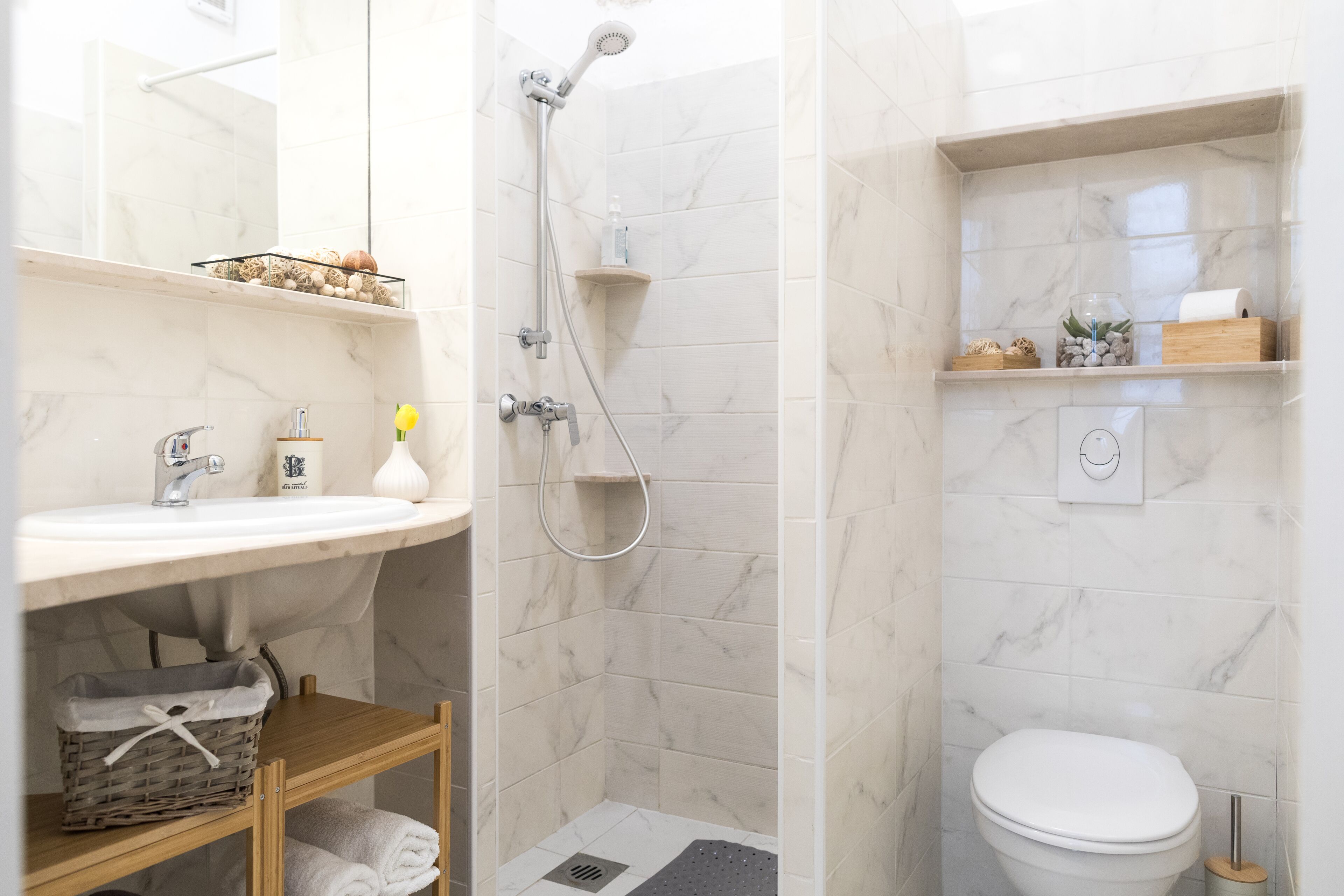 A bathroom with a white sink, a shower, and a toilet. There are shelves with toiletries and a basket under the sink.