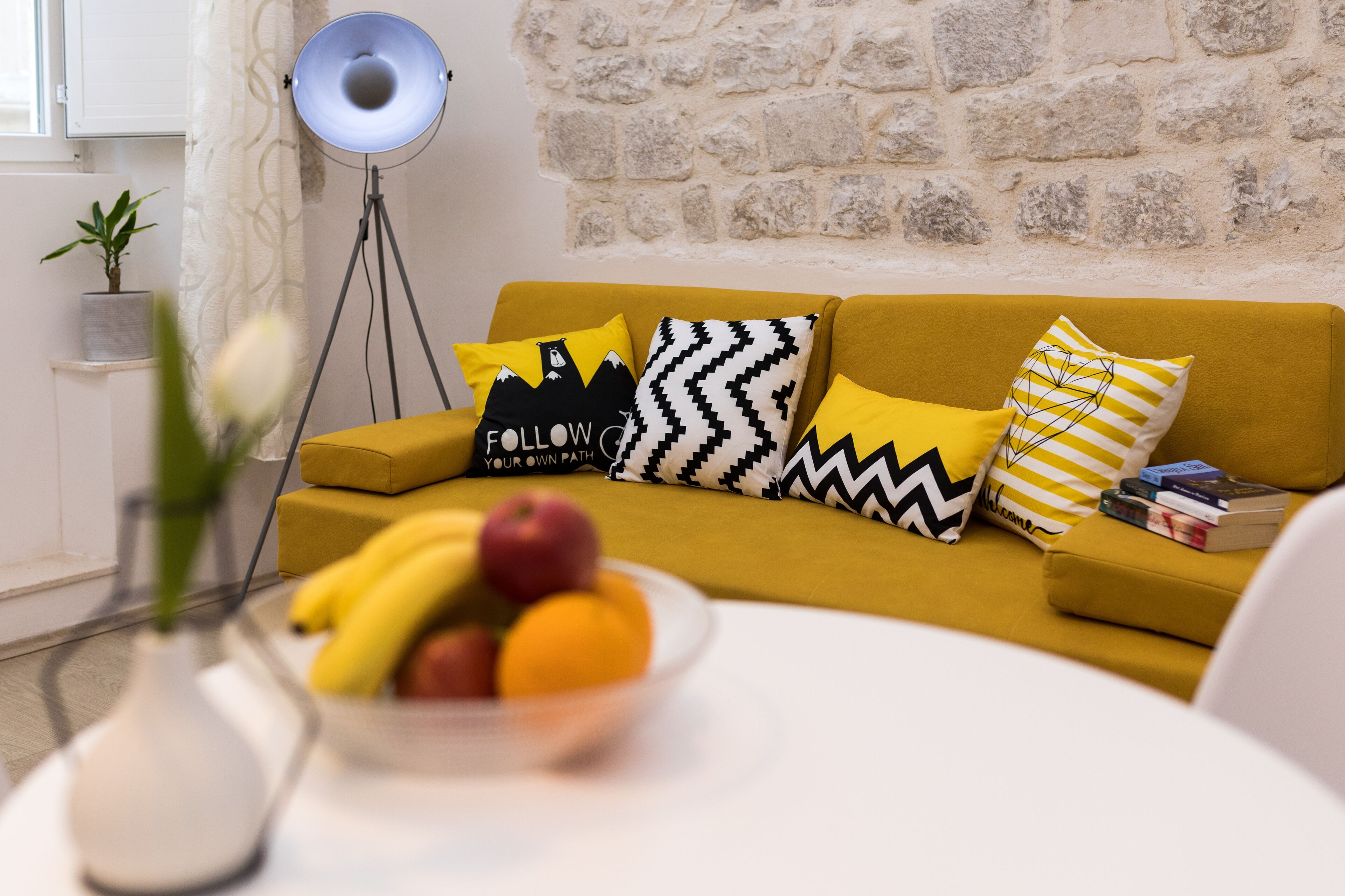A living room with a mustard sofa, patterned cushions, a floor lamp, a small table with a fruit bowl, and a potted plant.