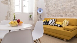A dining area with a white table and chairs, a bowl of fruit, and a yellow sofa with patterned cushions. - Apartment Toni (Dubrovnik)