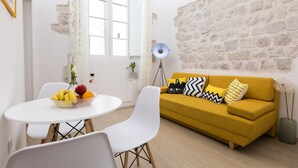 A modern living room with a yellow sofa, white chairs, a round dining table with fruit, and a stone wall. - Apartment Toni (Dubrovnik)