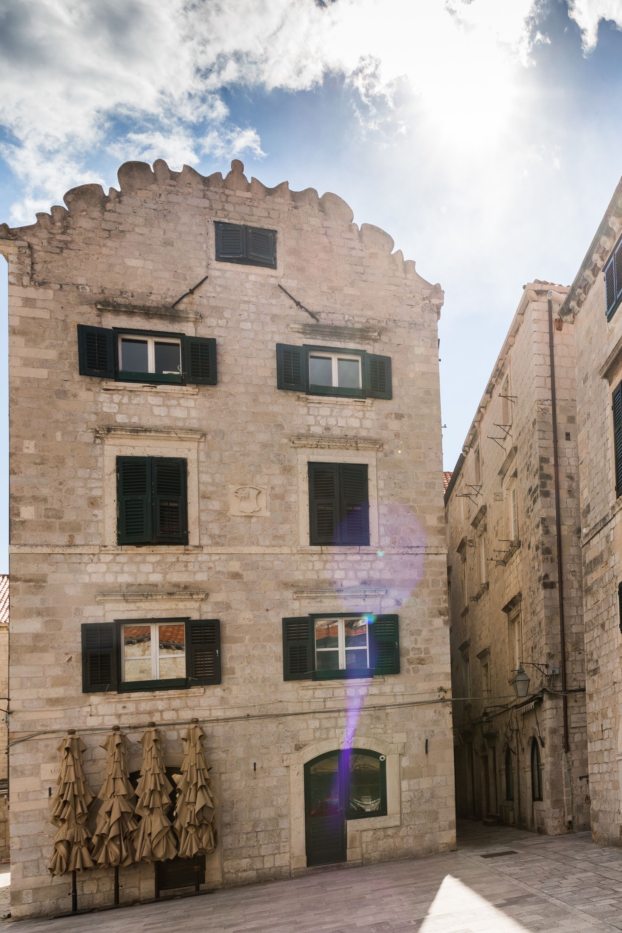 A historic stone building with multiple windows and shutters, a narrow alleyway, and a row of umbrellas.
