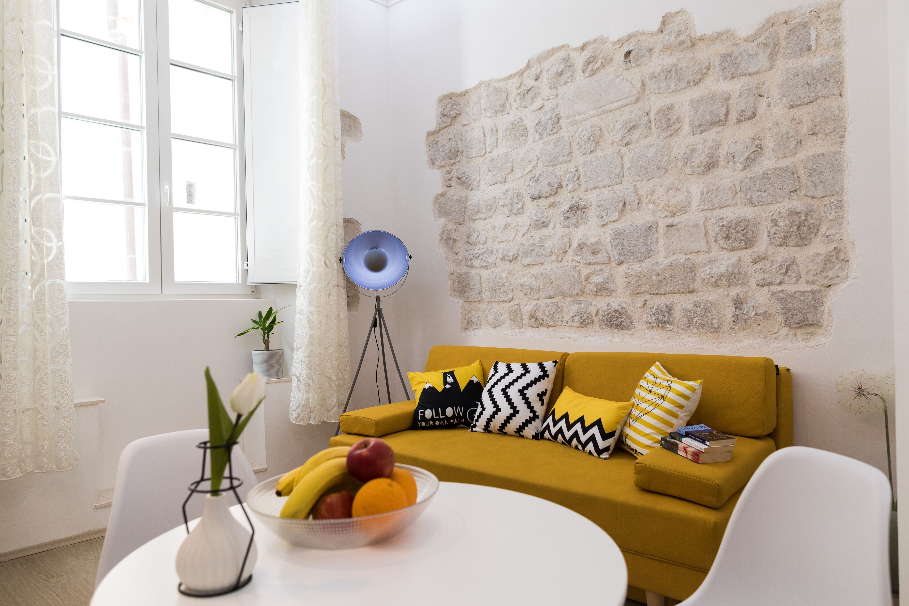 A room with a mustard-colored sofa, a white table with a fruit bowl, a potted plant, and a lamp.