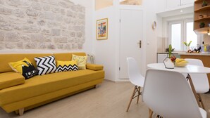 A modern living room with a yellow sofa, white chairs, a small dining table, and a kitchen area with a window. - Apartment Toni (Dubrovnik)