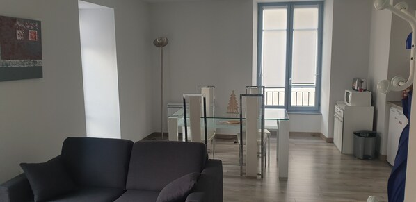 Una sala moderna con sofá, comedor con mesa de vidrio y cocina con electrodomésticos blancos.