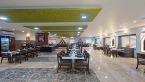 A spacious restaurant with a long central table, multiple seating areas, and a modern ceiling design. - Hotel Kaushal International (Sanchor)