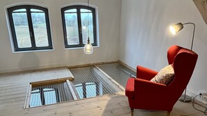 Ein Raum mit hohen Decken und schrägem Dach, zwei Fenstern, einem roten Sessel, einer Hängeleuchte und Holzboden.
