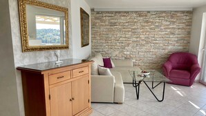 Un salon avec un mur en pierre, un meuble en bois, un canapé beige, une table basse en verre et un fauteuil violet.