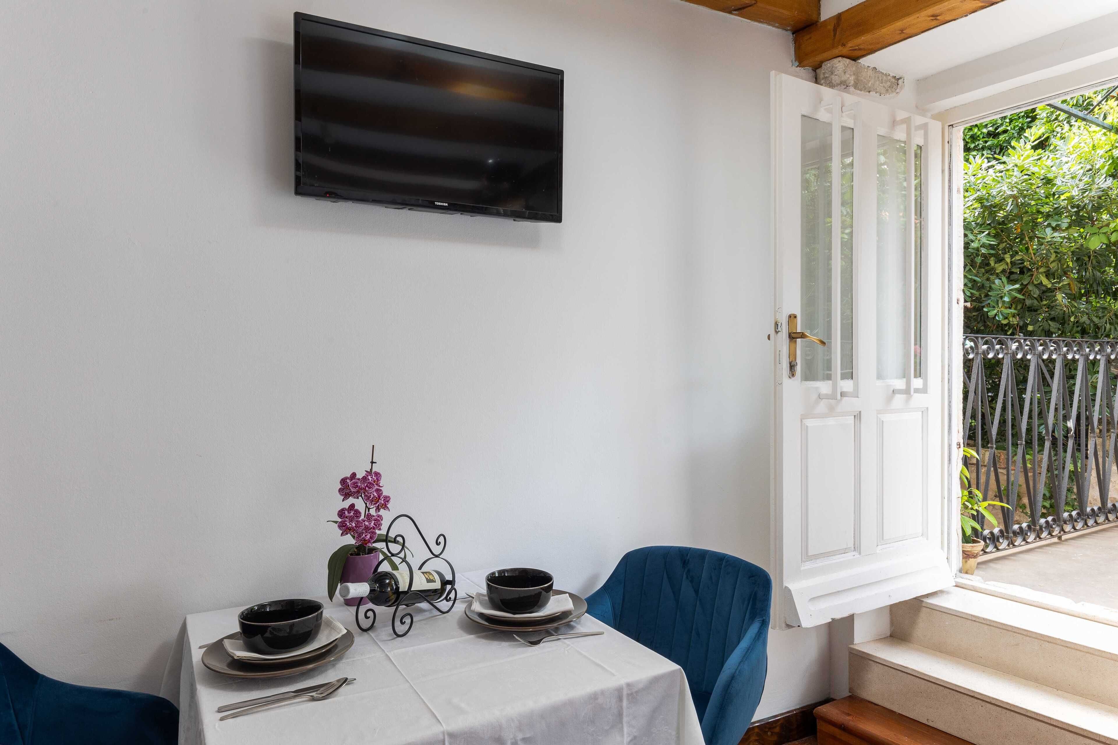 Un coin repas avec une table dressée pour deux personnes, un téléviseur fixé au mur et une porte donnant sur un balcon extérieur.