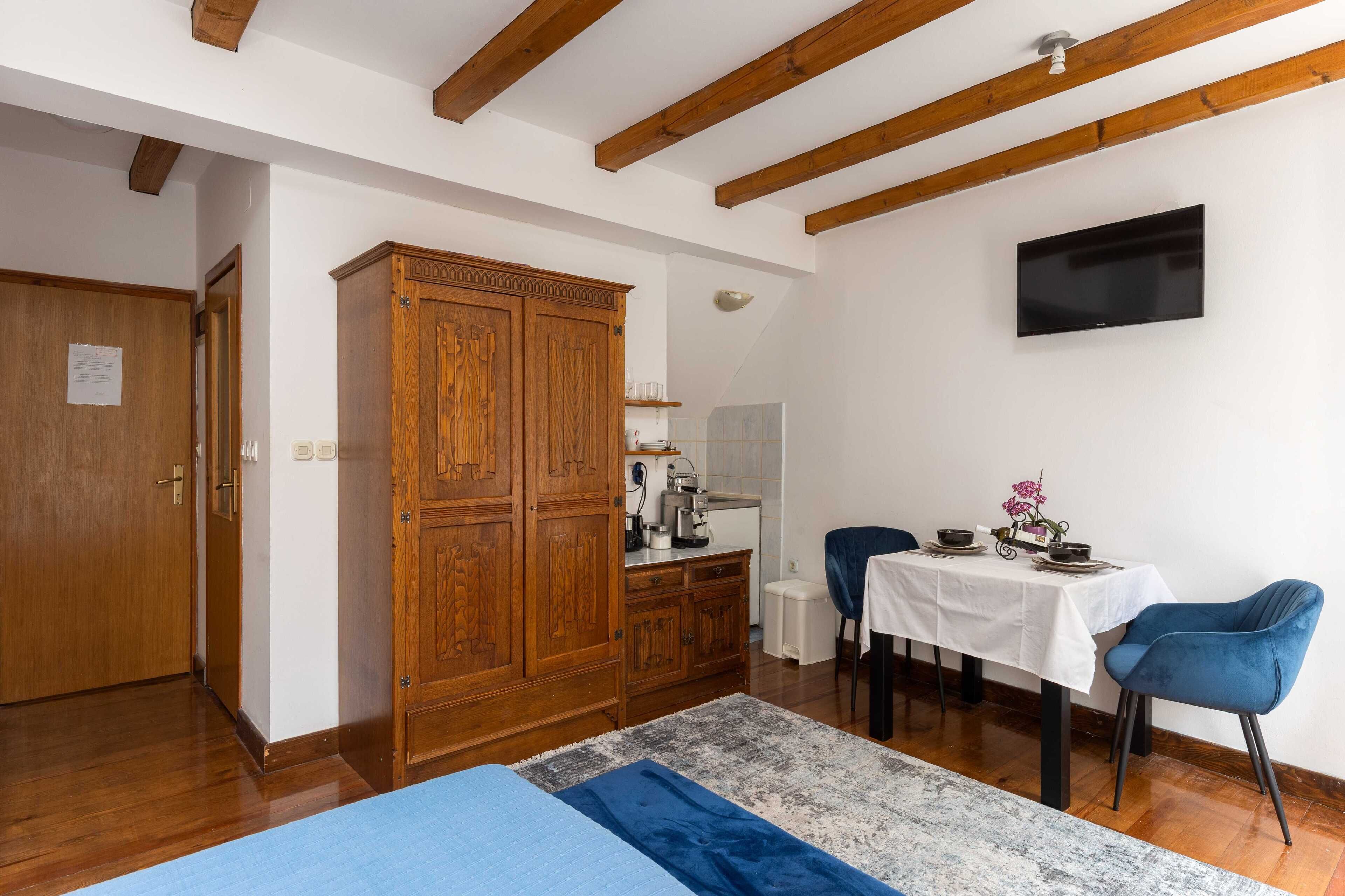 A room with wooden beams, a TV mounted on the wall, a dining table with chairs, and a cabinet.