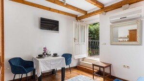 A room with a wooden ceiling, a white wall, a television, a table with chairs, a small table, a mirror, and a window with a view of greenery. - Apartment Taura (Dubrovnik)