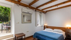 A bedroom with a wooden ceiling, a bed with a blue bedspread, a bedside table, a nightstand with a lamp, and a window with curtains. - Apartment Taura (Dubrovnik)