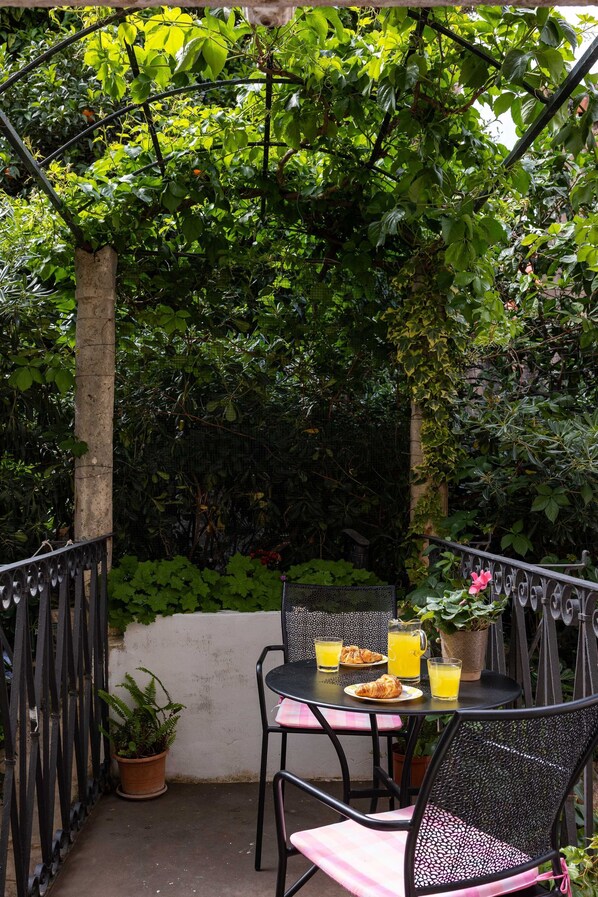 Un patio al aire libre con una mesa dispuesta para el desayuno, incluyendo croissants y zumo de naranja, rodeado de abundante vegetación y una valla de metal.