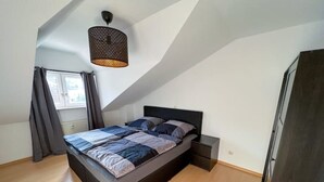 Quarto moderno com teto inclinado, cama com cabeceira escura, mesa de cabeceira e luminária pendente.