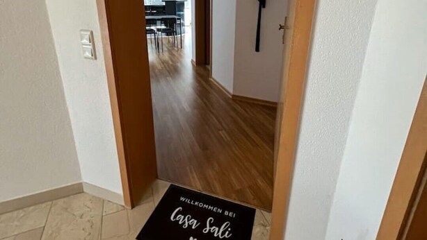 Um corredor com um tapete preto com a inscrição "Willkommen bei Casa Salis", piso de madeira e um vaso de planta.