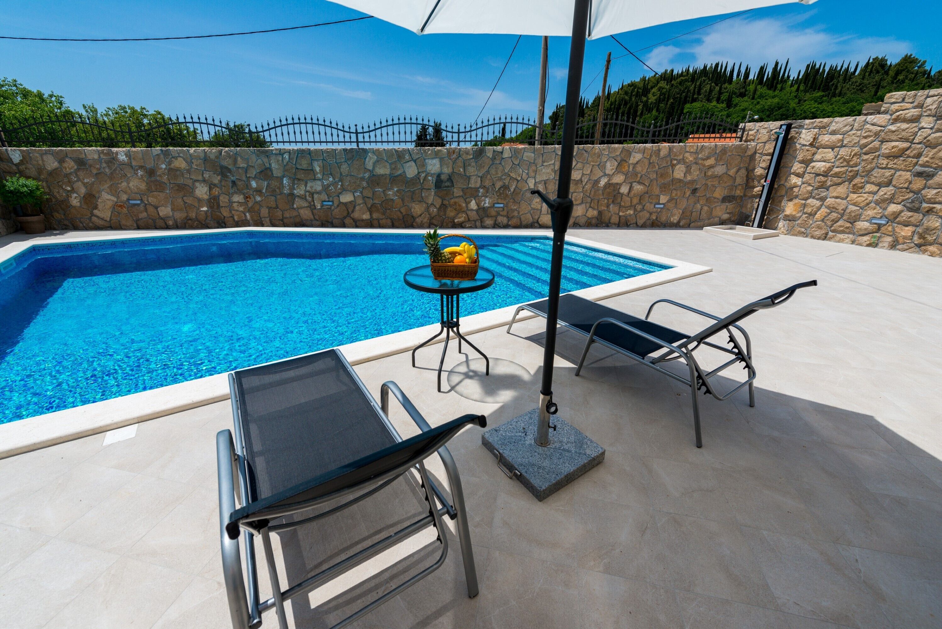 Una piscina con sillas de descanso y una mesa, rodeada de muros de piedra y una valla.