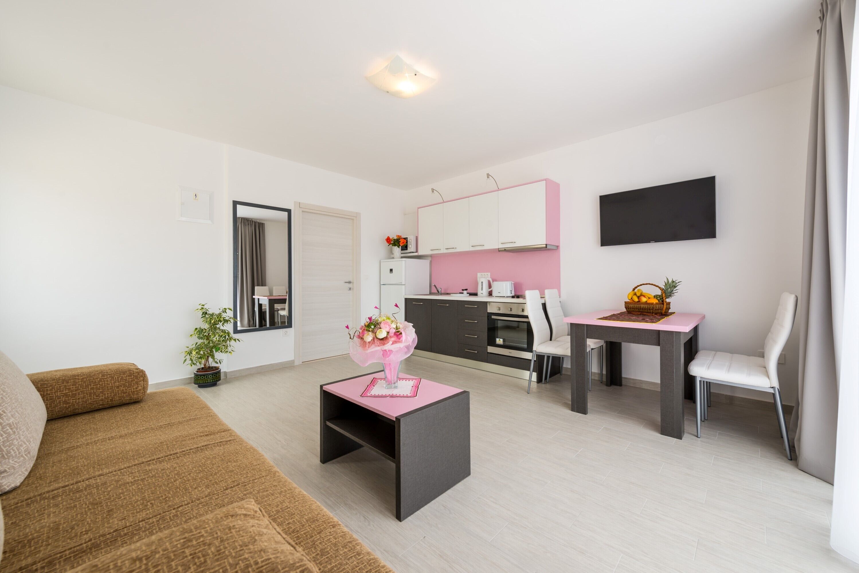 A modern living room with a sofa, a small coffee table, a dining area with a table and chairs, a kitchen with pink cabinets, and a flat-screen TV mounted on the wall.