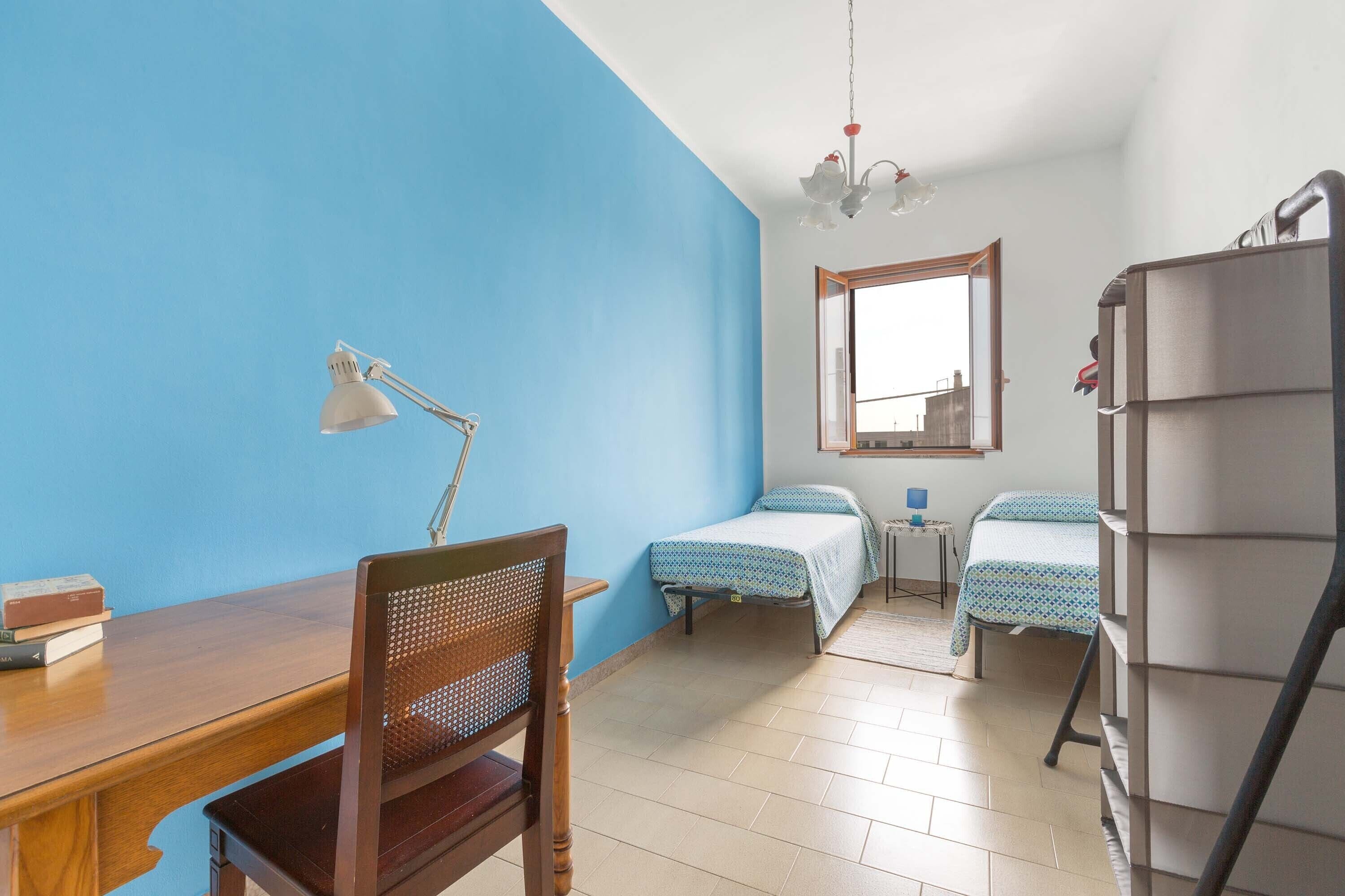 A room with a wooden desk, two single beds, a chair, a small table, and a bookshelf.