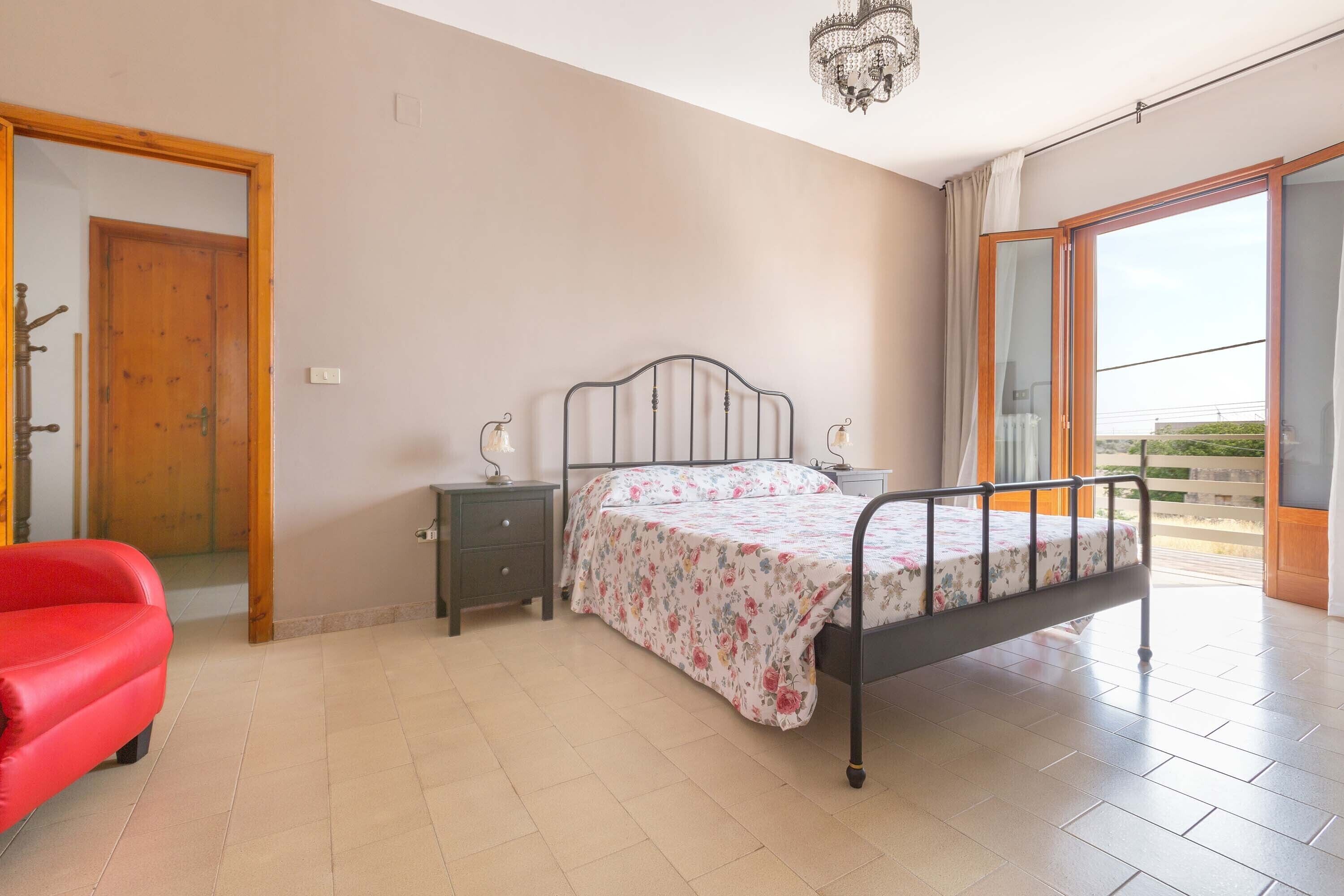 Une chambre à coucher avec un lit, une table de chevet, une chaise rouge, une porte en bois et une grande fenêtre.