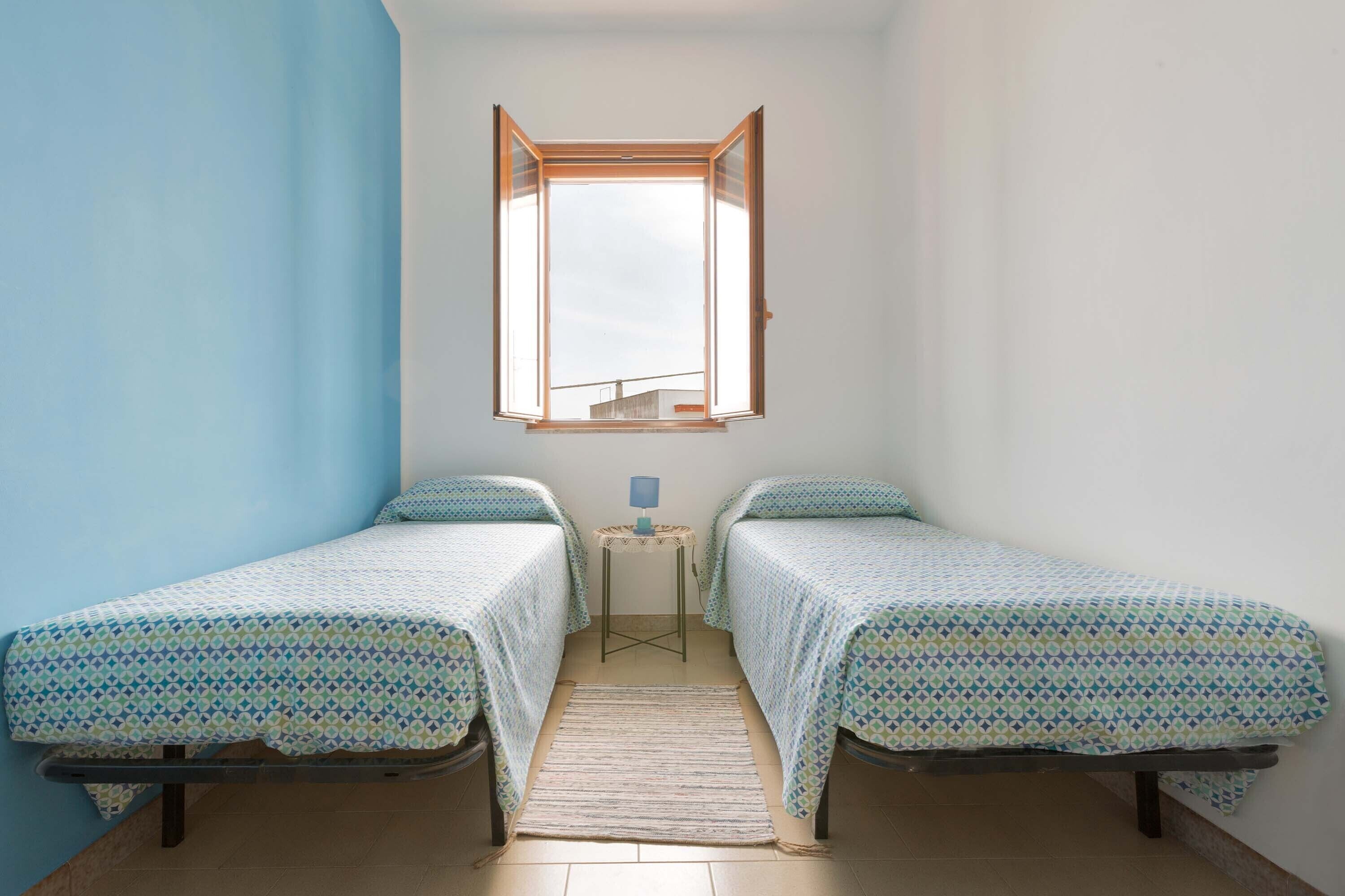 Quarto pequeno com duas camas de solteiro, uma mesinha com abajur e uma janela com a persiana aberta.