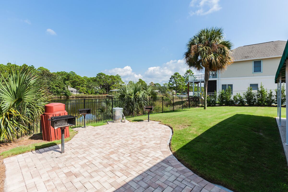 A paved patio area with a grill, trash bins, and a fenced yard with a lawn and palm tree.
