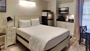 A bedroom with a bed, bedside table, lamp, ceiling fan, and a kitchenette with a microwave.
