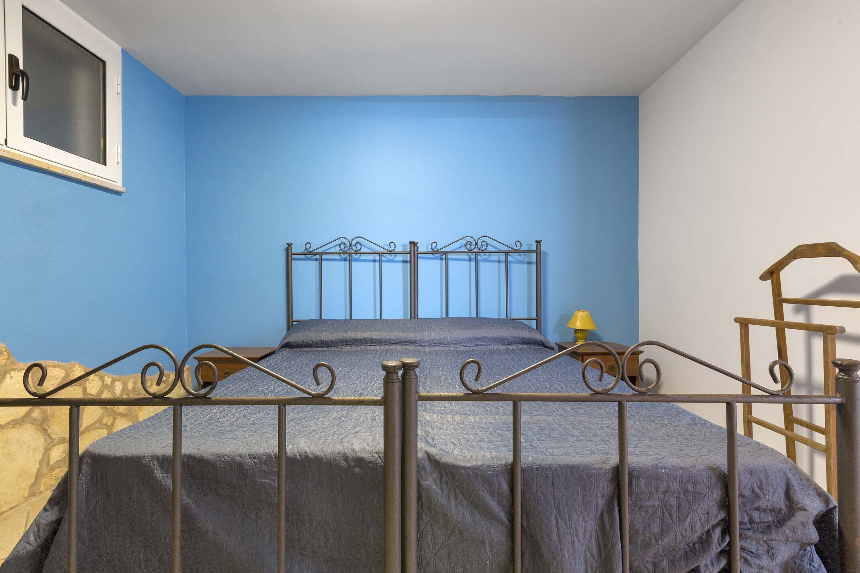 Uma cama de solteiro com estrutura metálica e cabeceira, uma janela com cortina e uma escada de madeira encostada na parede.