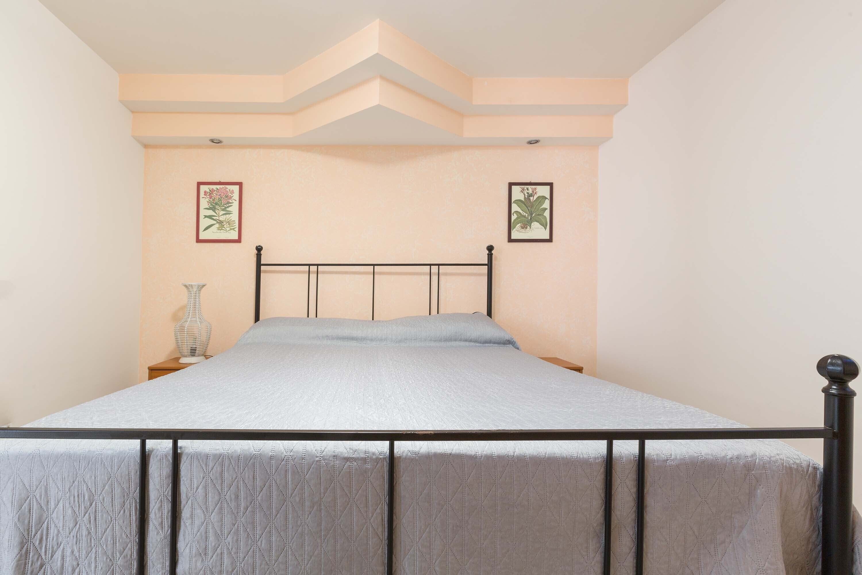 A bedroom with a bed, a nightstand, a vase, and two framed botanical prints on the wall.
