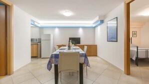 A compact dining area with a table set for four, a television, and a kitchenette in the background. - 2703 Villino Mare Blu by Barbarhouse (Melendugno)