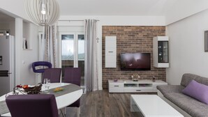A modern living room with a TV mounted on a brick wall, a dining area with purple chairs, and a white sofa with a purple pillow. - Apartment H&D (Dubrovnik)