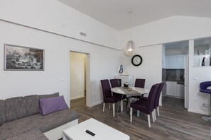 A modern living room with a gray sofa, purple cushions, a dining table with chairs, a kitchen in the background, and a clock on the wall.