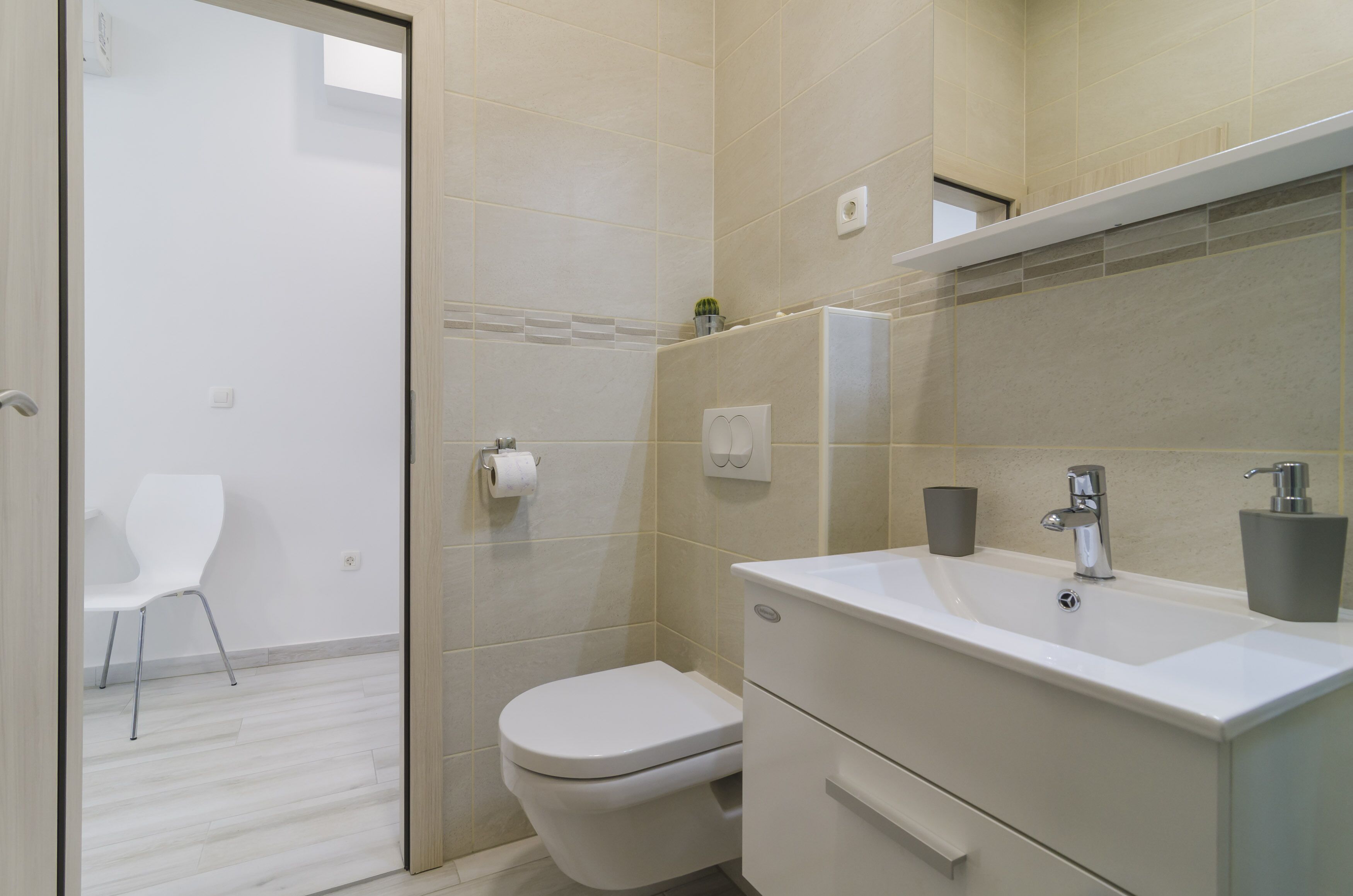 A modern bathroom with a toilet, sink, and mirror. There is a chair in the adjacent room.