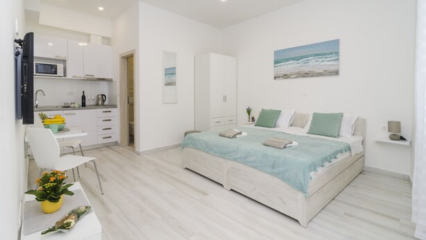 A modern bedroom with a bed, a desk, a chair, a small kitchen area, and a bathroom. - Apartment Rafaello (Dubrovnik)