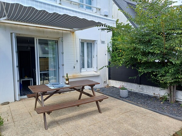 A patio with a wooden table and chairs, a bottle of champagne, and a small table with a glass.