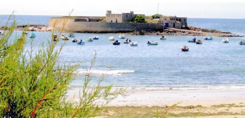 Paisaje costero con barcos, una construcción fortificada y una playa.