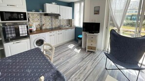 A compact kitchen with white cabinets, a microwave, oven, and a washing machine. There's a dining area with a table and chairs, and a TV on a stand.
