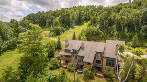 A large, two-story house with a prominent front porch, surrounded by dense forest and a sloping hill. - Ski In / Ski Out Treetops Home with Outdoor Private Spa (Stratton)