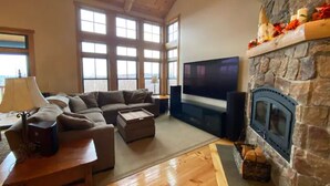 A spacious living room with a stone fireplace, a large TV, and a sofa. - Ski In / Ski Out Treetops Home with Outdoor Private Spa (Stratton)