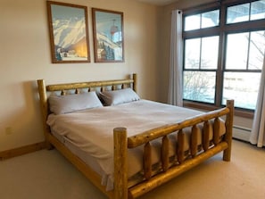 A wooden bed with a headboard, a window with curtains, and two paintings on the wall.