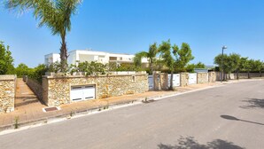 A residential street with a stone wall, a palm tree, and a white building with multiple garage doors. - 3046 Villetta Bianca by Barbarhouse (Otranto)