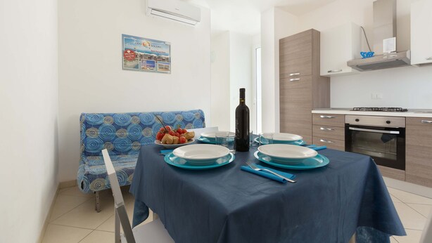 A dining area with a table set for four, a blue sofa, and a kitchen with modern appliances. - 2371 Villette Girovagando - Trilo Superior by Barbarhouse (Melendugno)