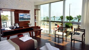 A hotel room with a bed, a desk, a chair, and a large window offering a view of the beach and palm trees. - UK Blue Coast Hotel  - Adults Only (Alanya)