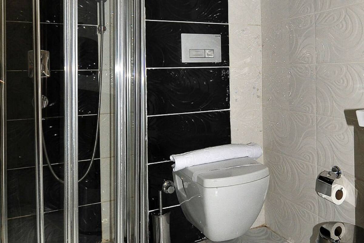 Une salle de bain avec une douche à parois de verre, des toilettes, une poubelle et un téléphone fixé au mur.