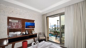 A hotel room with a bed, a desk with a TV, a chair, a window with a view of a pool and palm trees, and a red trash bin. - UK Blue Coast Hotel  - Adults Only (Alanya)