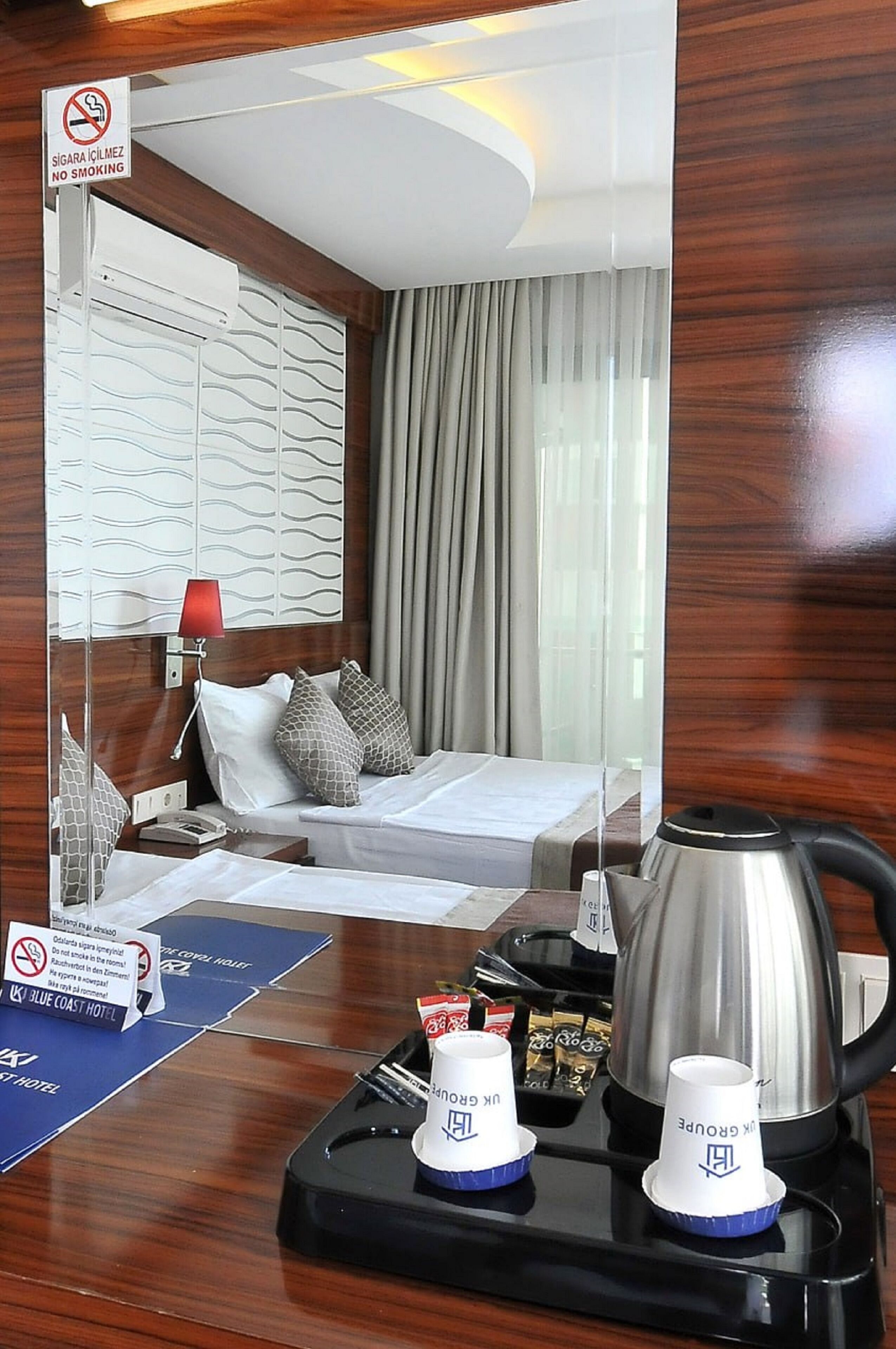 A hotel room with a bed, a desk with a kettle, a tray with cups, and a no-smoking sign.