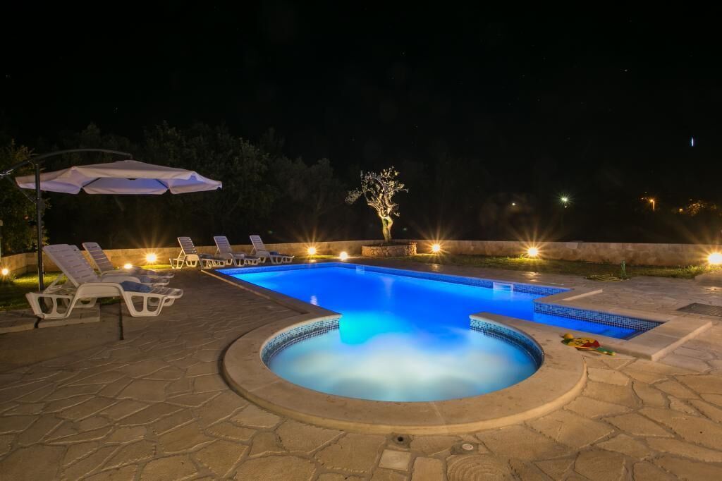 Una piscina con sillones de descanso y un jacuzzi, rodeada de baldosas de piedra e iluminación nocturna.
