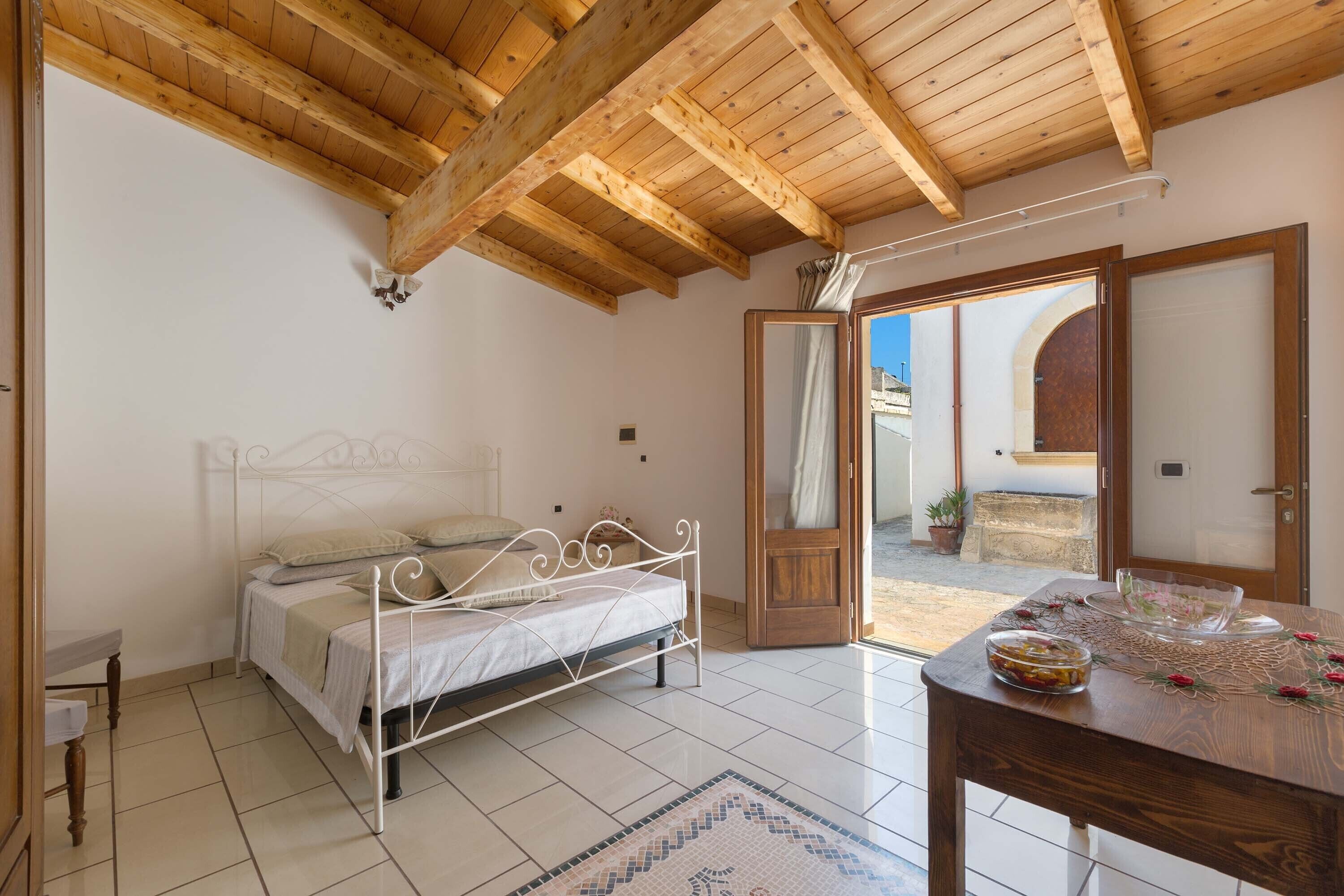 Quarto com teto de madeira, uma cama, uma mesa com uma tigela e vista através de uma porta aberta.